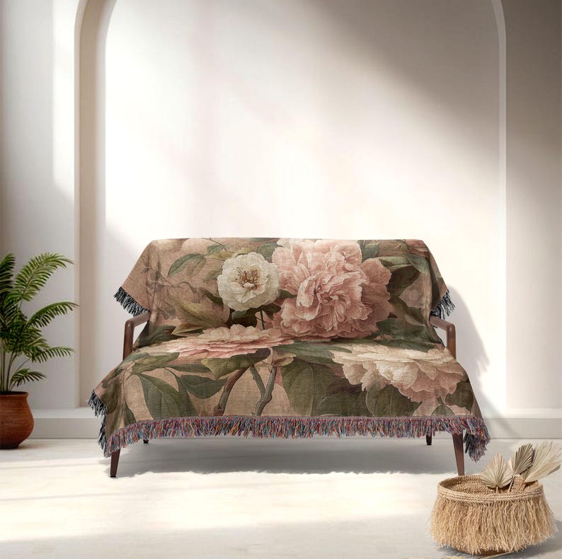 May include: A floral throw blanket on a wooden sofa. The blanket displays large pink and cream peony flowers with green leaves on a beige backdrop. The edges have a fringe trim.
