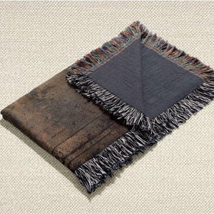 May include: A folded brown and black throw blanket with a distressed, textured appearance. The blanket has a dark gray underside and is trimmed with a fringe of black, white, and multicolored threads. The blanket is displayed on a cream-colored, textured surface.