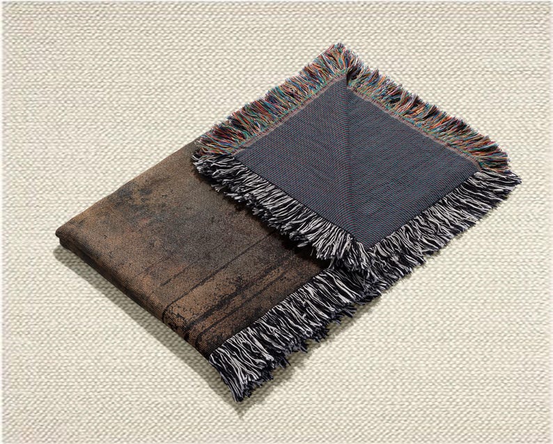 May include: A folded brown and black throw blanket with a distressed, textured appearance. The blanket has a dark gray underside and is trimmed with a fringe of black, white, and multicolored threads. The blanket is displayed on a cream-colored, textured surface.
