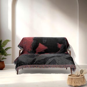 May include: A woven throw blanket with a black gorilla design on a red and grey backdrop. The blanket is draped over a wooden sofa, with a fringe along the bottom edge. The gorilla graphic is detailed and prominent.