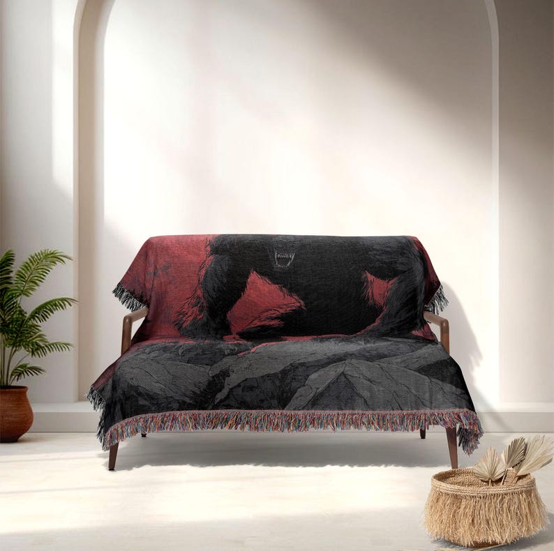 May include: A woven throw blanket with a black gorilla design on a red and grey backdrop. The blanket is draped over a wooden sofa, with a fringe along the bottom edge. The gorilla graphic is detailed and prominent.