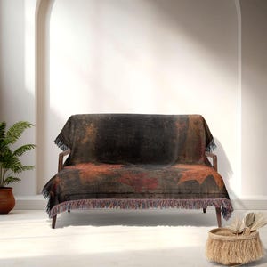 May include: A dark-colored throw blanket with a leaf pattern draped over a wooden sofa. The blanket features shades of black, brown, and red, with a fringed edge. The sofa is set against a white wall.