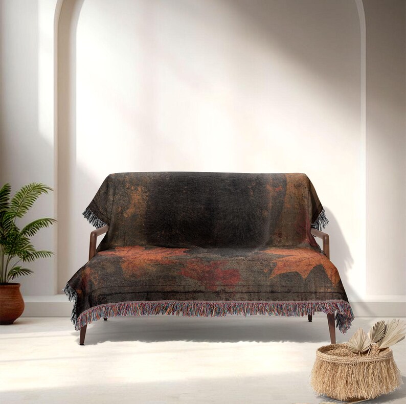May include: A dark-colored throw blanket with a leaf pattern draped over a wooden sofa. The blanket features shades of black, brown, and red, with a fringed edge. The sofa is set against a white wall.