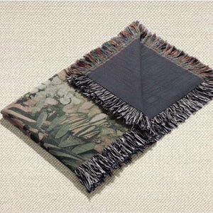 May include: Folded woven throw blanket with a botanical print in shades of green, brown, and pink. The blanket has a dark grey backing and fringed edges with a mix of colours. The blanket is folded on a textured, off-white surface.