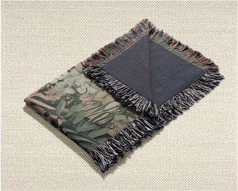 May include: Folded woven throw blanket with a botanical print in shades of green, brown, and pink. The blanket has a dark grey backing and fringed edges with a mix of colours. The blanket is folded on a textured, off-white surface.