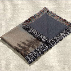 May include: A folded throw blanket with a landscape design featuring trees in shades of brown and gray. The blanket has a dark gray underside and a fringed edge with a mix of colors. The blanket is on a textured, off-white surface.