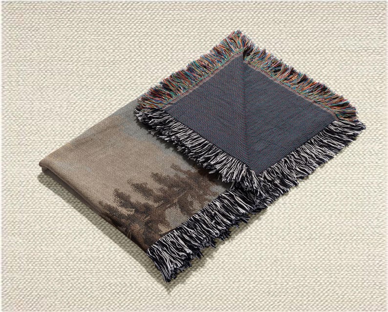 May include: A folded throw blanket with a landscape design featuring trees in shades of brown and gray. The blanket has a dark gray underside and a fringed edge with a mix of colors. The blanket is on a textured, off-white surface.