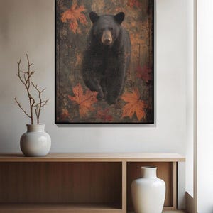 May include: A framed art print featuring a black bear walking forward, set against a backdrop of autumn leaves. The bear has dark fur and a light brown snout. The leaves are shades of orange and red, creating a rustic, nature-inspired aesthetic.