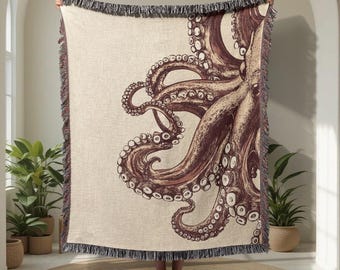 May include: A beige throw blanket with a detailed illustration of an octopus in brown tones. The blanket has a fringed edge. The octopus design is the focal point, with its tentacles prominently displayed. The blanket is held up.