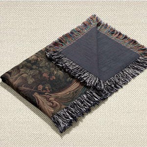 May include: Folded tapestry throw blanket with a dark green and brown floral pattern. The blanket has a dark gray backing and fringed edges with a mix of colors. The blanket is folded on a light beige textured surface.