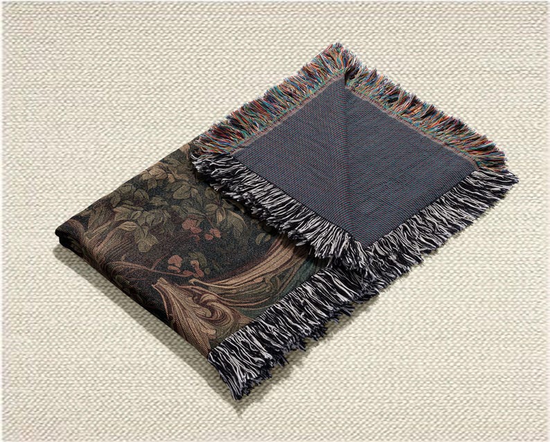 May include: Folded tapestry throw blanket with a dark green and brown floral pattern. The blanket has a dark gray backing and fringed edges with a mix of colors. The blanket is folded on a light beige textured surface.