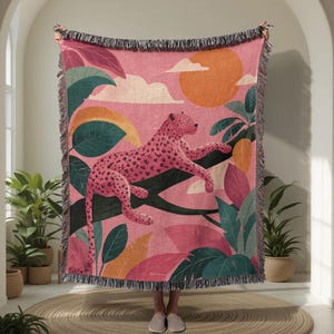 May include: A pink woven throw blanket featuring a leopard resting on a tree branch, surrounded by colorful leaves and a setting sun. The blanket has a fringed edge and is held up by a person.