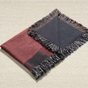 May include: A folded burgundy throw blanket with a dark grey section and fringed edges. The fringe includes black, white, and multicoloured threads. The blanket is displayed on a textured, off-white surface.