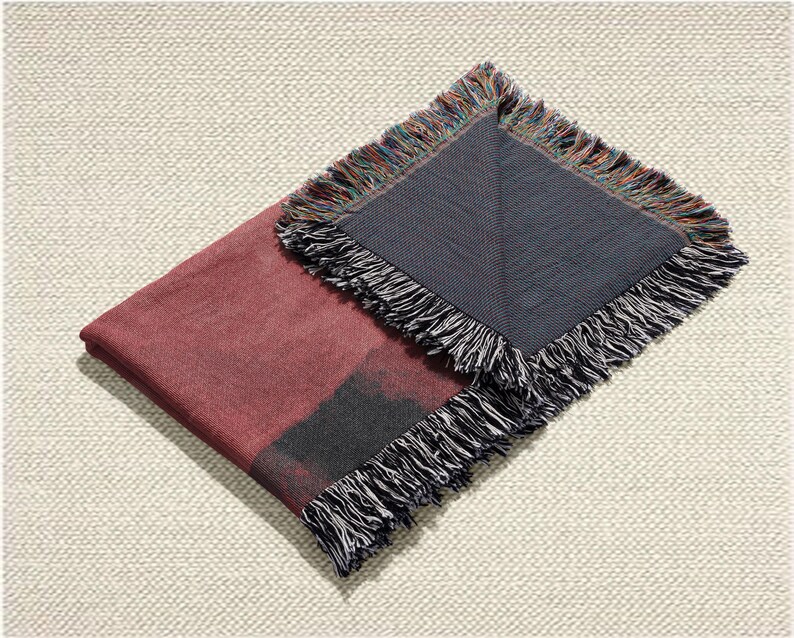 May include: A folded burgundy throw blanket with a dark grey section and fringed edges. The fringe includes black, white, and multicoloured threads. The blanket is displayed on a textured, off-white surface.