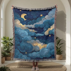 May include: A woven throw blanket featuring a night sky design with a crescent moon, stars, and swirling clouds in shades of blue, gold, and cream. The blanket has a fringed edge and is held up by a person.