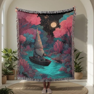 May include: A woven throw blanket with a fantasy illustration of a gnome in a boat on a river, surrounded by a forest at night. The blanket has a fringed edge and a colour palette of pink, teal, and black.