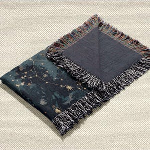 May include: A folded throw blanket with a dark teal and navy blue celestial design featuring constellations and stars. The blanket has a fringed edge with a mix of colours, and the reverse side is a solid dark grey.
