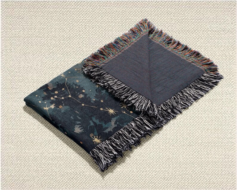 May include: A folded throw blanket with a dark teal and navy blue celestial design featuring constellations and stars. The blanket has a fringed edge with a mix of colours, and the reverse side is a solid dark grey.