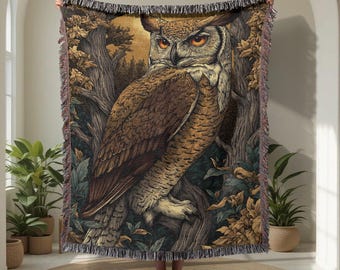May include: A woven throw blanket featuring a detailed illustration of a large owl with orange eyes perched on a tree branch. The design includes a forest background with golden-brown leaves and dark tree trunks, finished with a fringed edge.
