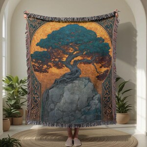 Peut inclure: Plaid tissé avec une illustration détaillée d'un arbre sur un rocher. Le motif comprend des tons sarcelle, marron et or, avec une bordure décorative et des franges. La couverture est tenue, montrant son design complet.