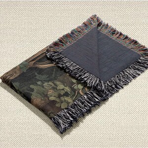 May include: Folded tapestry throw blanket with a dark forest scene featuring trees and foliage in shades of green, brown, and black. The blanket has a fringed edge with a mix of colors. The underside is a solid dark gray.