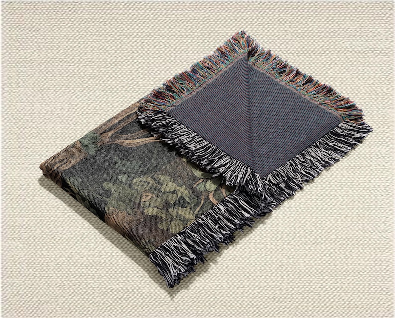 May include: Folded tapestry throw blanket with a dark forest scene featuring trees and foliage in shades of green, brown, and black. The blanket has a fringed edge with a mix of colors. The underside is a solid dark gray.
