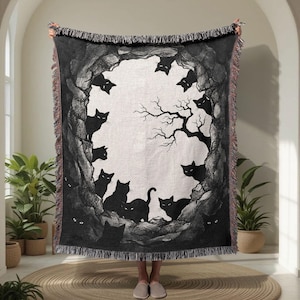 Könnte beinhalten: Eine gewebte Wohndecke mit einer schwarz-weißen Illustration mehrerer schwarzer Katzen, die aus einer felsigen Höhlenöffnung hervorschauen. Das Design zeigt eine kahle Baumsilhouette vor hellem Hintergrund und einen dekorativen Fransenrand.