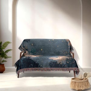May include: A dark blue throw blanket with a celestial design featuring stars and a forest scene. The blanket is draped over a wooden sofa and has a fringed edge. The colour palette includes shades of blue, gold, and brown.