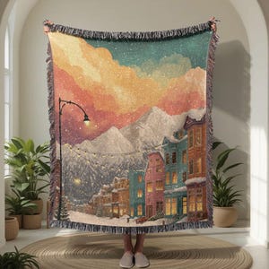 Könnte beinhalten: Eine gewebte Decke mit einer Winterszene mit schneebedeckten Bergen, farbenfrohen Gebäuden und einer Straßenlaterne. Die Decke hat einen Fransenrand und zeigt eine verschneite Stadt unter einem Sonnenuntergangshimmel. Die Farbpalette umfasst Blau-, Orange- und Grüntöne.