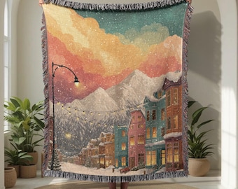 May include: A woven throw blanket featuring a winter scene with snow-covered mountains, colorful buildings, and a street lamp. The blanket has a fringed edge and depicts a snowy town under a sunset sky. The color palette includes blues, oranges, and greens.