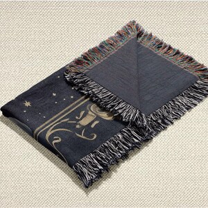 May include: A dark blue throw blanket with a celestial design featuring stars and a decorative border. The blanket has a fringed edge with a mix of colors. The design includes a stylized illustration in a light tan color.