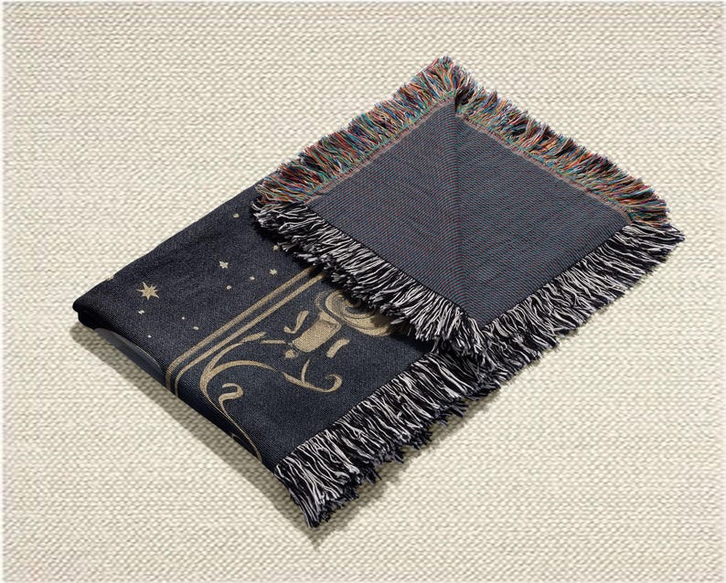 May include: A dark blue throw blanket with a celestial design featuring stars and a decorative border. The blanket has a fringed edge with a mix of colors. The design includes a stylized illustration in a light tan color.
