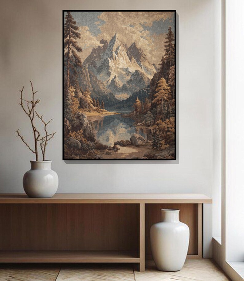 May include: A framed landscape painting depicts a mountain range with snow-capped peaks reflected in a calm lake. The artwork features a color palette of blues, browns, and greens, with trees and clouds adding depth. The painting is displayed above a wooden console.