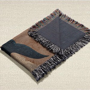 May include: A folded woven throw blanket with a brown and blue design. The blanket features a dark blue wing-like shape against a brown background with swirling patterns. The edges are finished with a fringe in multiple colors.