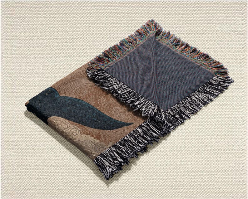 May include: A folded woven throw blanket with a brown and blue design. The blanket features a dark blue wing-like shape against a brown background with swirling patterns. The edges are finished with a fringe in multiple colors.