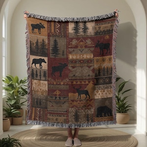 Pode incluir: Um cobertor tecido com um design de patchwork com silhuetas de alces, árvores e ursos em tons de castanho, bege e bordô. O cobertor tem uma borda com franjas e um tema rústico e ao ar livre.