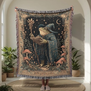 May include: A tapestry blanket featuring a frog wizard reading a book, wearing a blue robe and hat, and holding a staff. The design includes mushrooms, stars, and a crescent moon against a dark background. The blanket has a fringed edge.