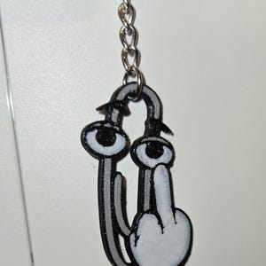 May include: A black and white 3D printed keychain featuring a stylized character with two large eyes on stalks and a hand gesture showing a middle finger. The figure is attached to a silver metal split ring by a short chain.