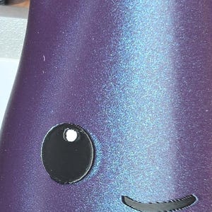 May include: A purple, iridescent, 3D-printed object with a smooth, metallic finish. It features a black, curved spout at the top, a single circular black eye with a white highlight, and a small, simple black curved line representing a mouth.