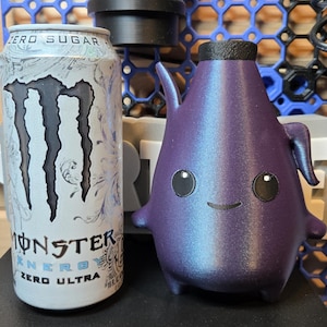 May include: A white Monster Energy Zero Ultra drink can next to a small, purple, pear-shaped figurine with a black cap, two small arms, two short legs, and a simple face featuring two round black eyes with white highlights and a thin black smile.