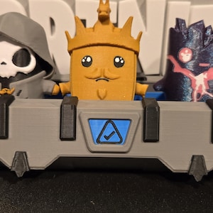 May include: Three small 3D-printed figurines displayed behind a gray and light blue rectangular barrier. The figures include a gray hooded skull character, a gold crown-wearing character with a mustache, and a dark purple textured creature.