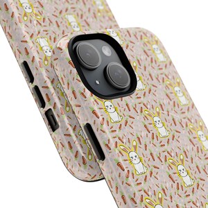 May include: A phone case with a repeating pattern of white bunnies and orange carrots on a light pink background. The case has a black camera lens and side buttons.