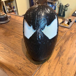 May include: A black mask with white eye coverings in a spider-like design. The mask is sitting on a wooden surface. The mask has a textured appearance.