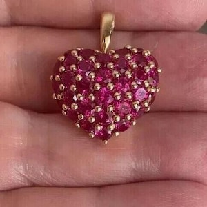 May include: A heart-shaped pendant with a gold bail, adorned with numerous small, vivid red gemstones. The gemstones are closely arranged, forming a textured surface. The pendant is a deep red hue, with gold detailing.