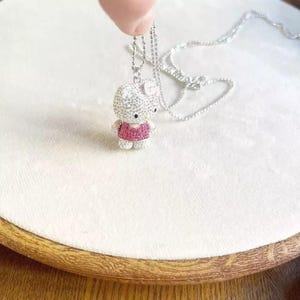 May include: A silver necklace featuring a sparkling, gem-encrusted pendant in the shape of a cartoon cat. The cat pendant is wearing a pink dress and bow. The necklace is displayed on a white surface.