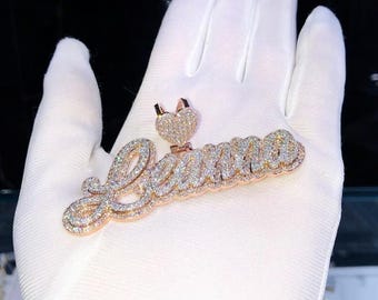 May include: A rose gold pendant spelling "Leanna" in a cursive font, encrusted with small, sparkling gemstones. A heart-shaped charm with a crown sits above the name. The pendant is displayed on a white surface.