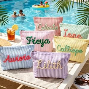 Op de afbeelding: Een verzameling gestreepte make-uptassen in verschillende kleuren, geborduurd met de namen Celeste, Freya, Ione, Calliope, Aurora en Lilith. De tassen worden getoond bij een zwembad, met een strohoed en ijsthee.