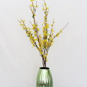 May include: A decorative arrangement featuring yellow forsythia branches with small flowers and green leaves in a green, ribbed glass vase. The vase has a metallic finish and a dark interior.