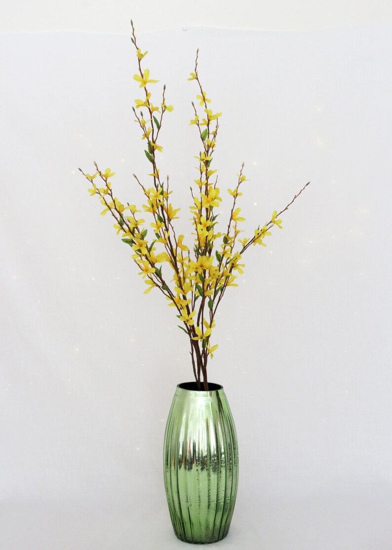May include: A decorative arrangement featuring yellow forsythia branches with small flowers and green leaves in a green, ribbed glass vase. The vase has a metallic finish and a dark interior.