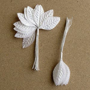 May include: Two white, artificial leaf stems. One stem features a cluster of seven leaves, while the other has a single leaf. The leaves have a textured, glittery appearance, and the stems are made of thin, white wire. These are likely for floral arrangements or craft projects.